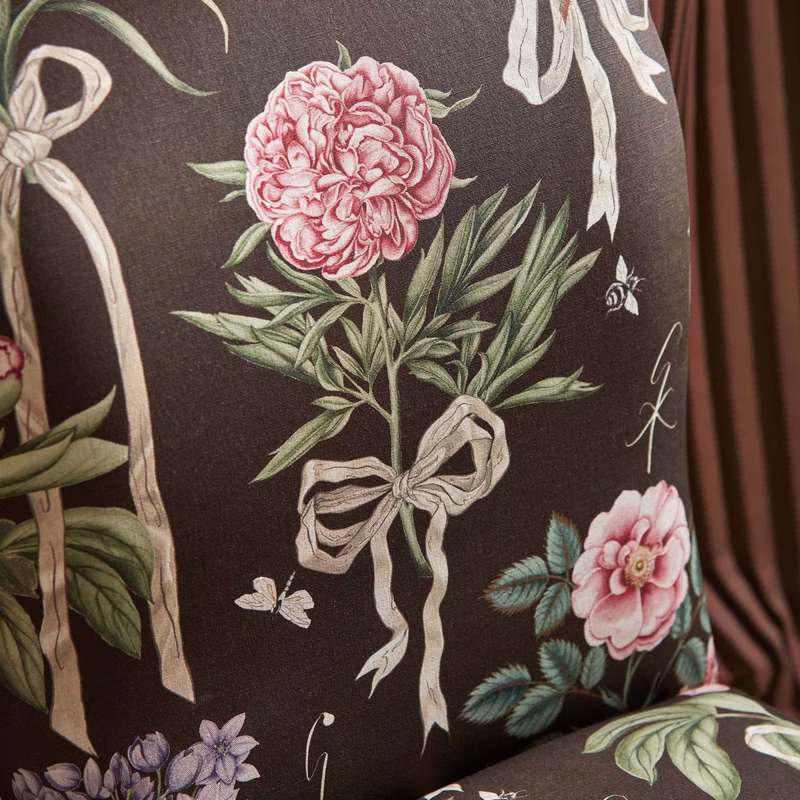 Floral printed upholstery fabric on a cushion, shown close up with pink flowers, leaves, ribbons and butterflies on a dark ground.