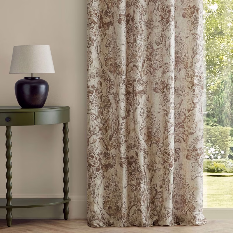 A room with a green side table and lamp beside patterned curtains made up in full-length drapes by a window.