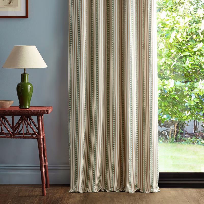 Room with a striped curtain hanging by a window, beside a table lamp and wicker table.