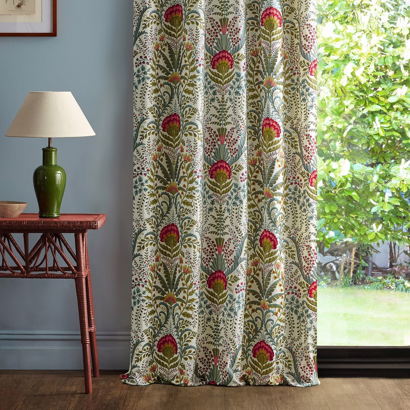 A room with printed Marrienne cloth made up as full-length curtains beside a window, with a table lamp and side table.