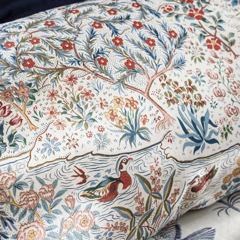 Patterned fabric with birds, flowers and trees is shown upholstered on a cushion, with another pale cushion beneath it.