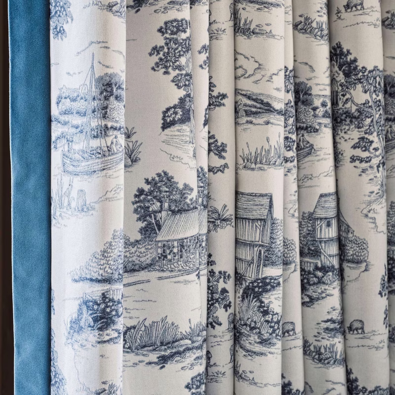 A close-up of patterned fabric hanging as curtains, showing a blue-and-cream countryside print with cottages, trees and riverside scenes.