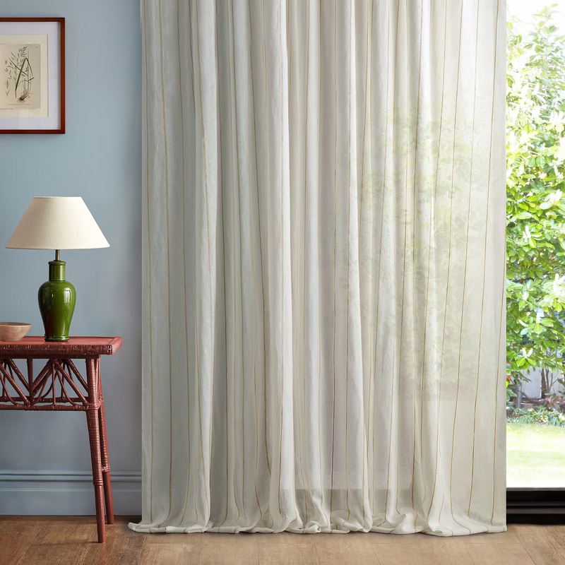 A room with floor-length sheer curtains hanging in soft vertical folds beside a small table and lamp, with the fabric showing a subtle pale striped pattern.