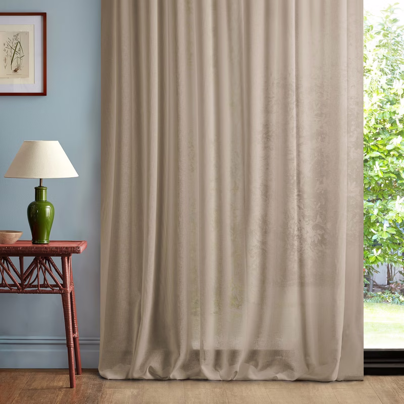 Interior room with a full-length sheer curtain hanging in soft folds by a window, with a table and lamp beside it.