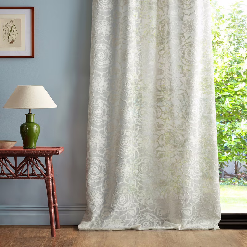 Light sheer patterned curtains hang floor-length beside a window in a room with a side table and lamp.