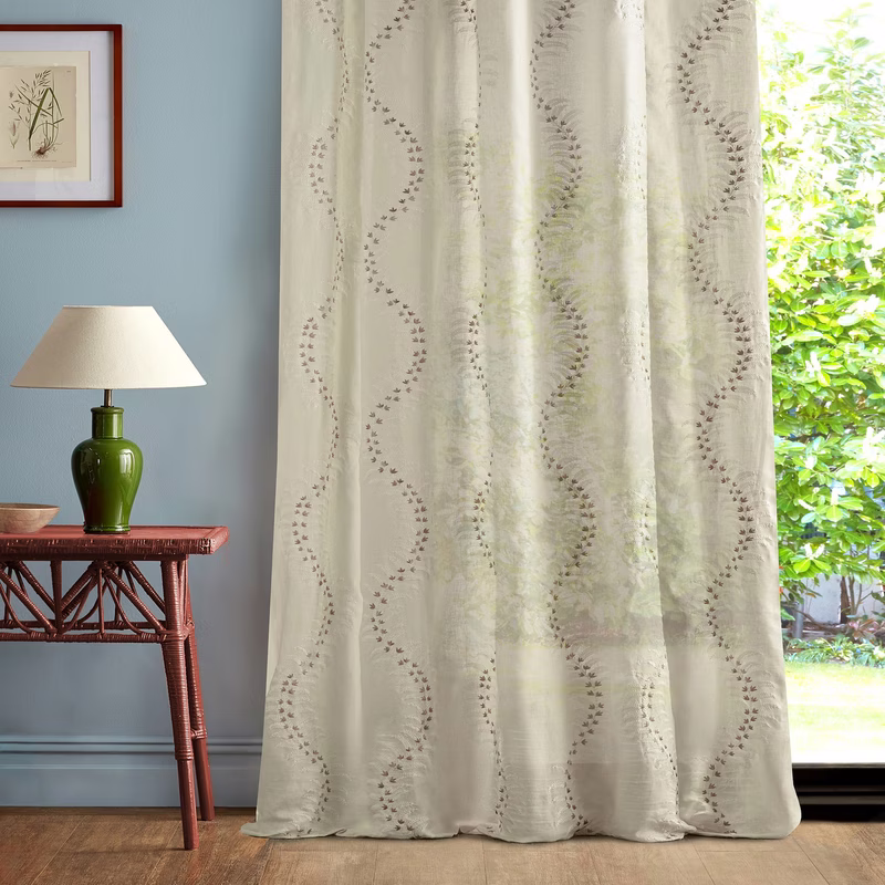 A room with a blue wall, side table and lamp, and sheer patterned curtains hanging at a window.
