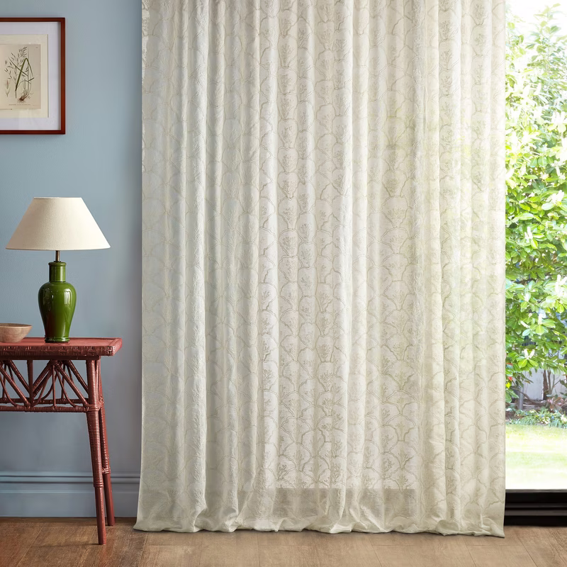 Light-patterned sheer curtains hang floor-length beside a window in a room with a lamp and side table.