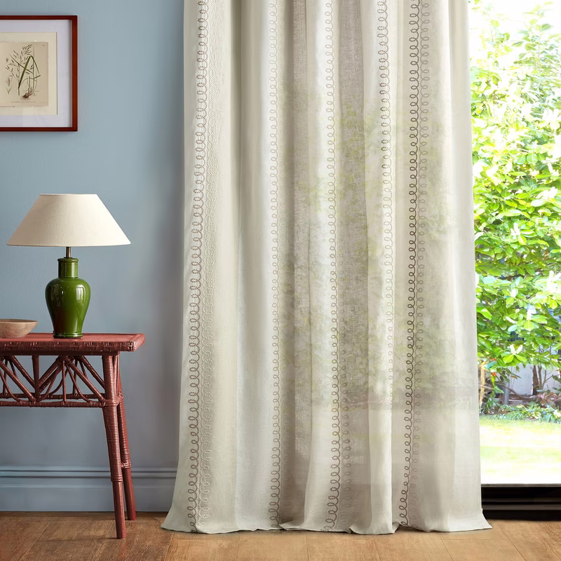Sheer curtains with a looped stripe pattern hang in a room beside a window, with a table lamp and side table visible.