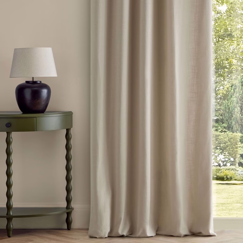 Neutral-toned room with floor-length curtains beside a window, and a lamp on a green side table.
