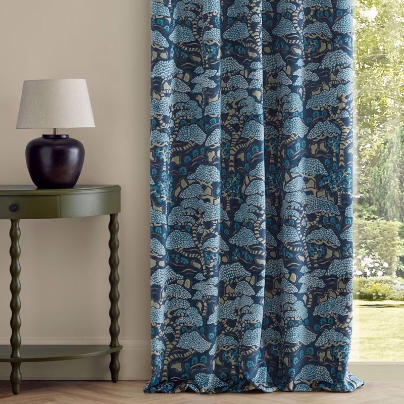 Room with a green side table and lamp beside curtains made from patterned Bonsai & Gingko fabric beside a window.