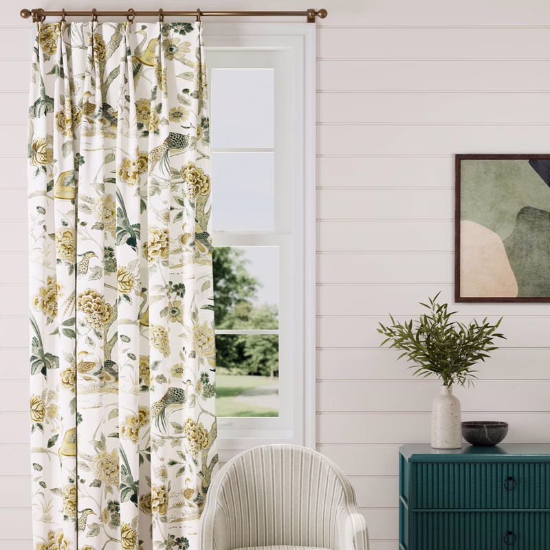 Curtains in a light floral and bird print hang at a window in a white-paneled room with a chair and sideboard.