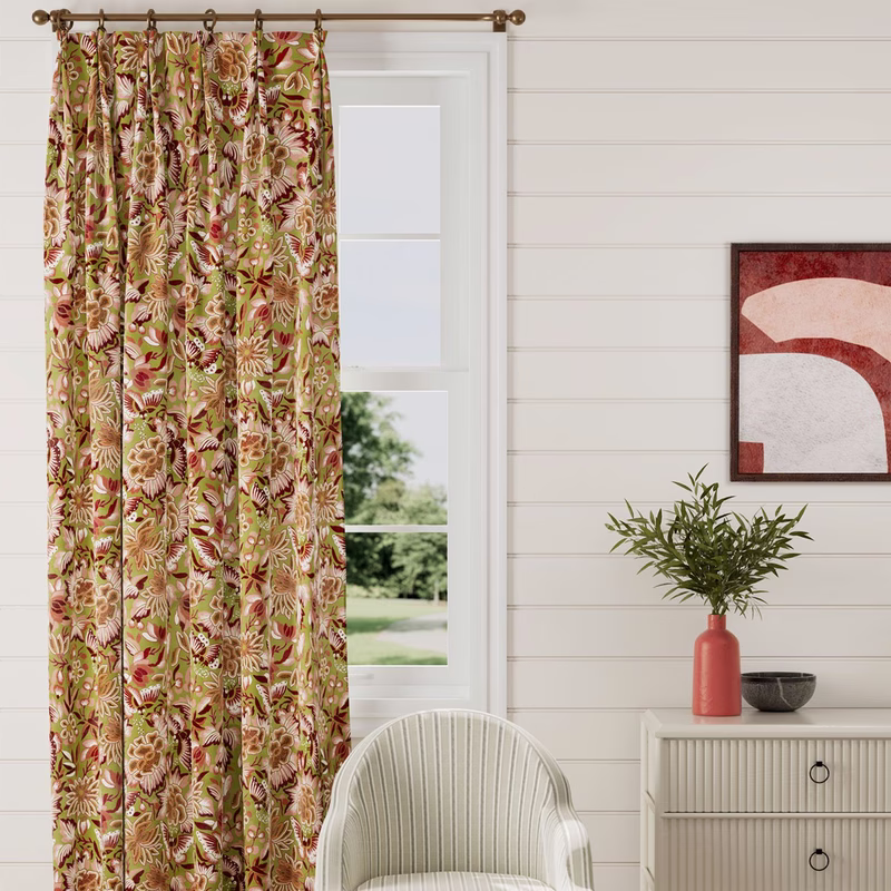 A bright room with a window dressed in full-length floral curtains on a brass pole, with cushions and furniture nearby.