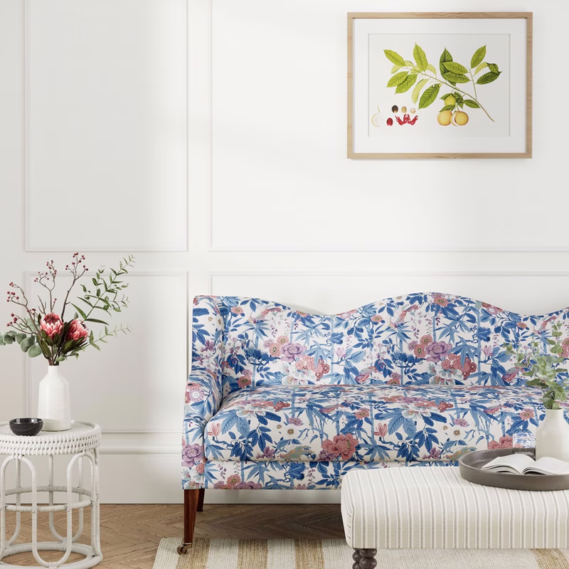 A floral blue-and-pink patterned fabric is upholstered on a sofa in a bright room with white walls and framed botanical prints.
