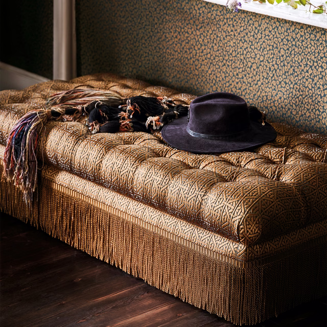 Bronze-gold geometric woven upholstery on a fringed bench with a black hat