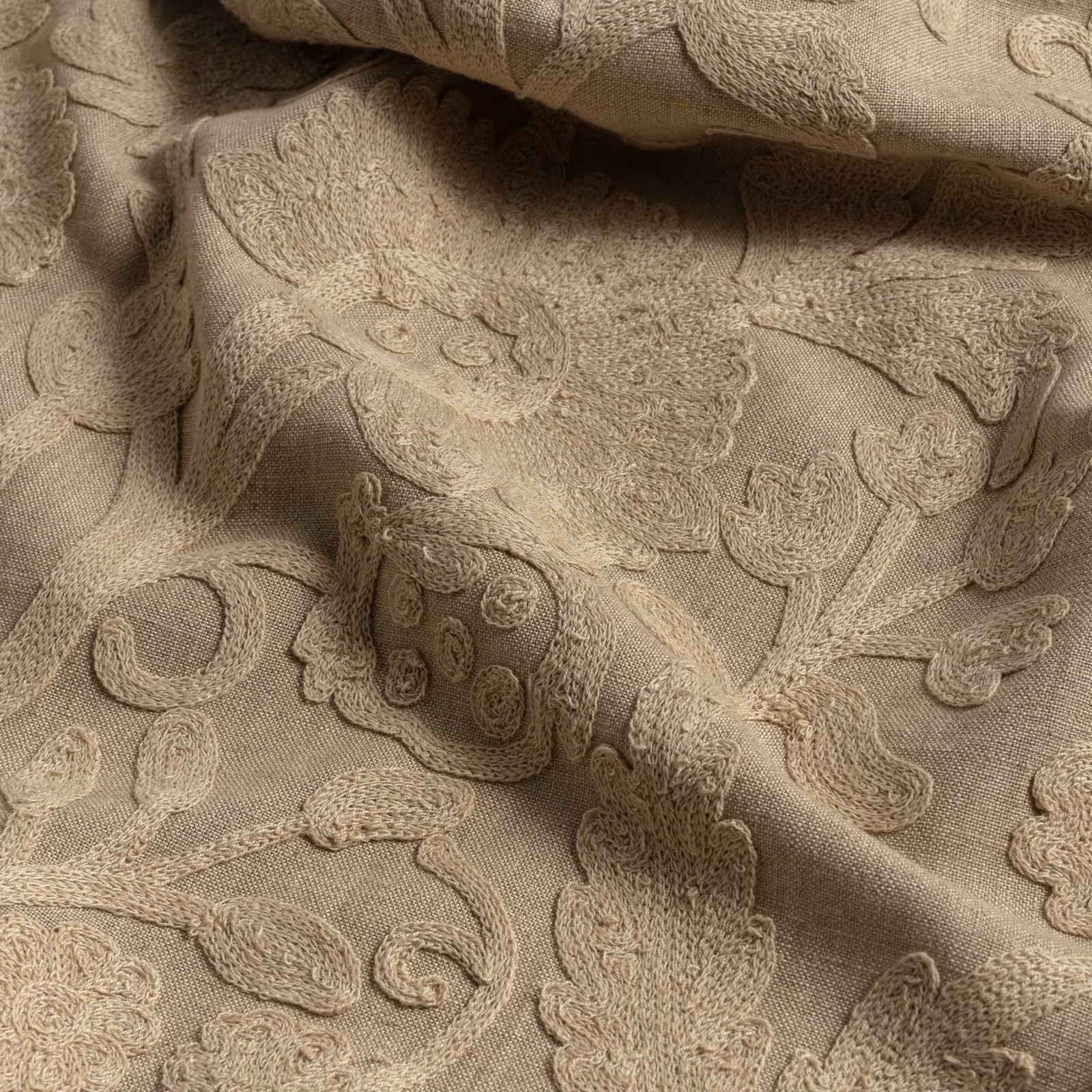 Warm flax-beige fabric with raised paisley, floral curls, and softly shadowed folds
